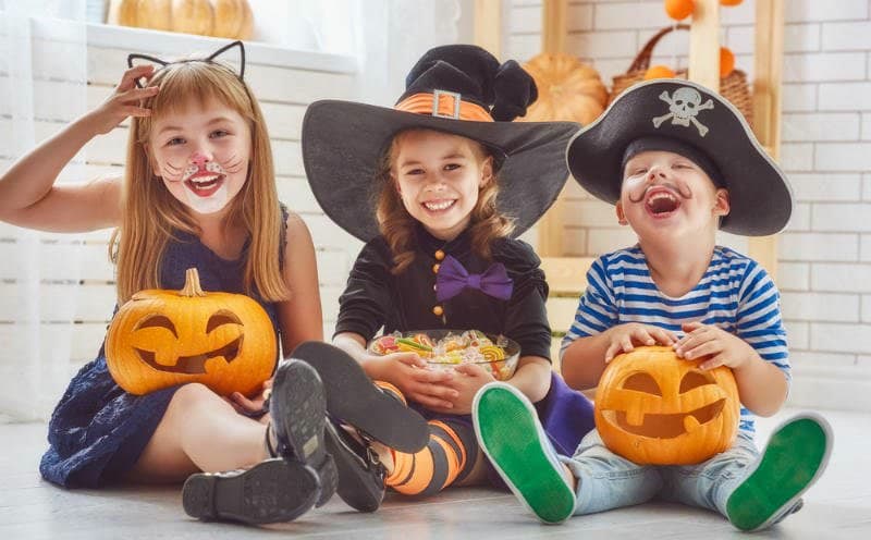 Three kidnergarten aged children dressed up as a cat, witch, and pirate sit on the floor with jack-o-lanters and candy in their laps. 