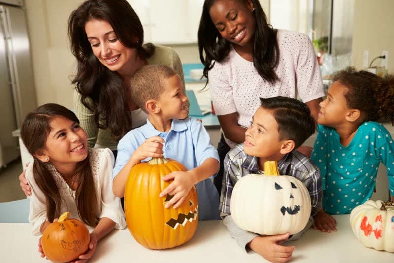 Two moms practice halloween safety tips while carving pumpkins at the kitchen counter with four children. 