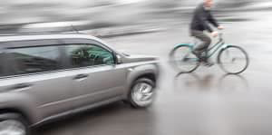 A silver car is about to hit a bicyclist on a rain drenched road.