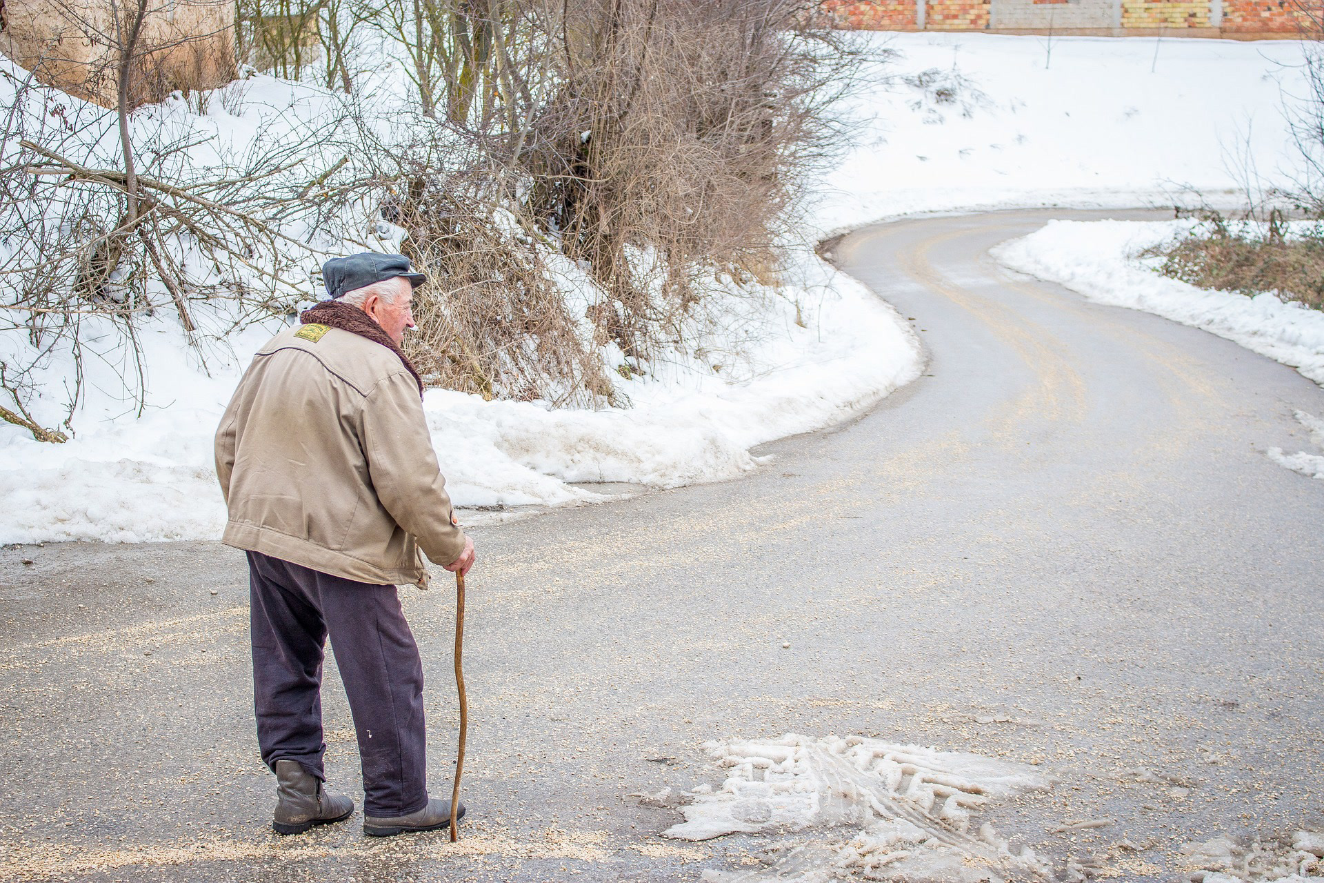 Elderly man with walking cane walks across icy road, which may result in a slip and fall injury.