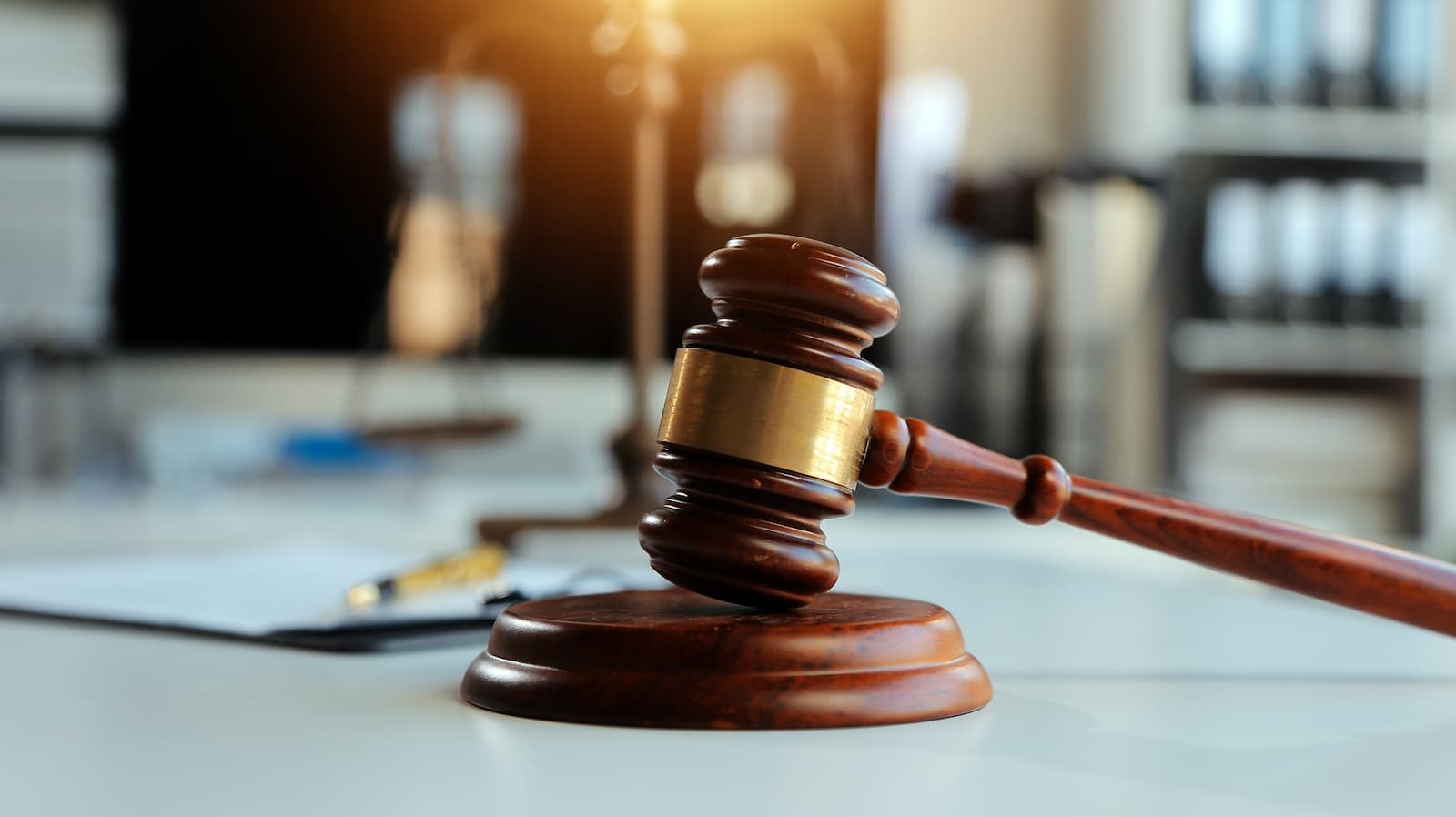 A close-up photo of a wooden gavel with a golden band on a light-colored desk in an office setting. The gavel is resting on its side on a round wooden base. In the background, a pen and a document are visible, along with blurred bookshelves and a scale of justice. The image is warmly lit, with a bright light source coming from the top left.