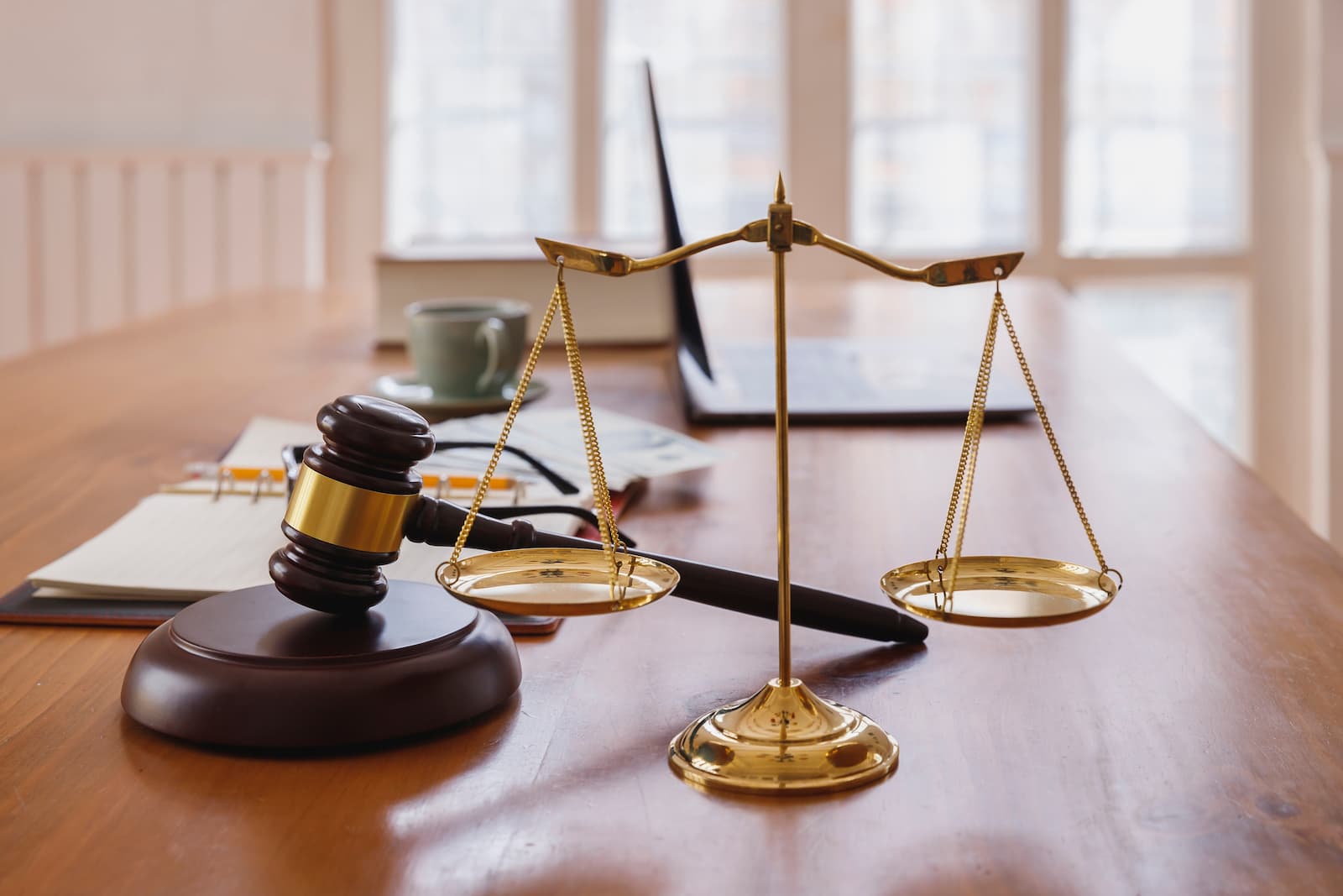 A low-angle photo of a wooden desk with two items in the foreground. On the left is a wooden gavel with a golden band, resting on a round wooden base. On the right is a small, golden scale of justice, with both pans empty and hanging level. In the background, a laptop, a cup of coffee, and some documents are visible on the desk, with a blurred window behind them.