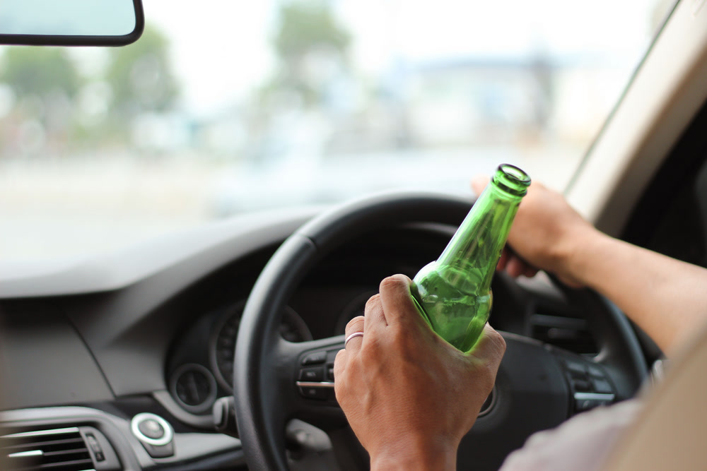 Man drives car in broad daylight with one hand on the steering wheel and the other holding a half full beer bottle.