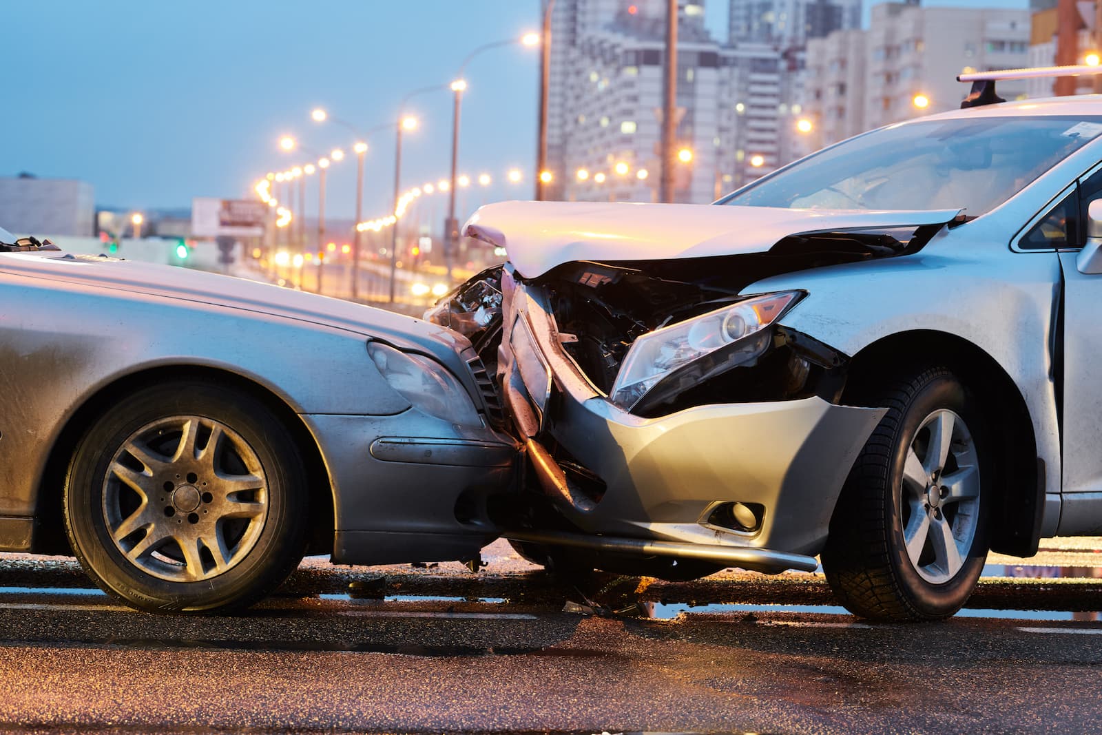 A professional photo of a head-on car collision between two silver cars at dusk. The front ends of both cars are severely crumpled, with shattered plastic and metal visible. The ground is wet and reflects the streetlights. The background is a blurry cityscape with streetlights and buildings.