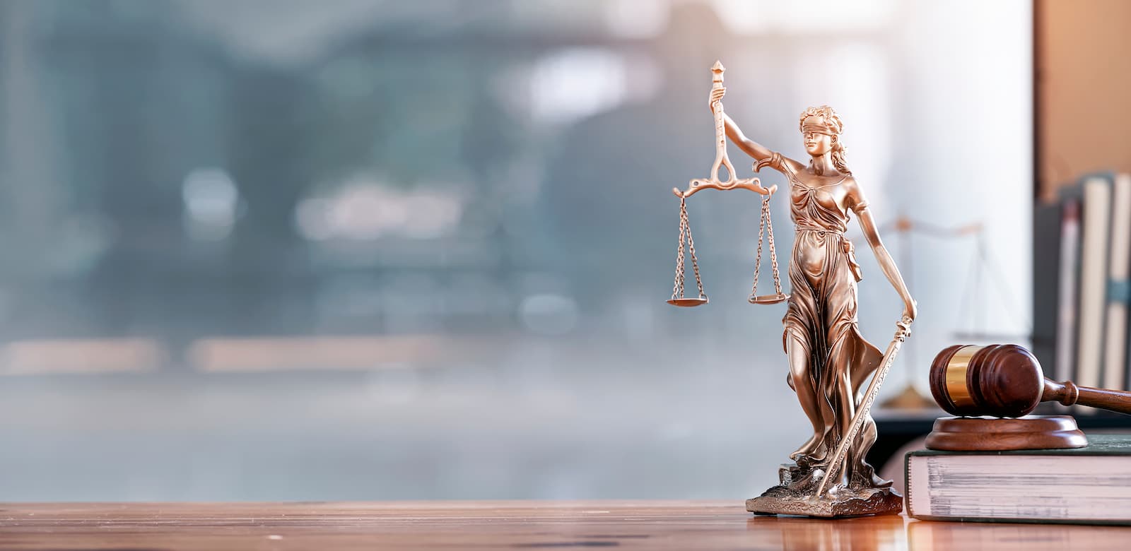 A wide, horizontal photo of a wooden desk with a bronze statue of Lady Justice and a gavel. The statue is in the foreground on the right, holding a scale in her left hand. The gavel is on a stack of legal books next to her. The background is a blurry office space with a large window and another scale of justice visible in the distance. The entire scene is lit with a warm light from the top right.