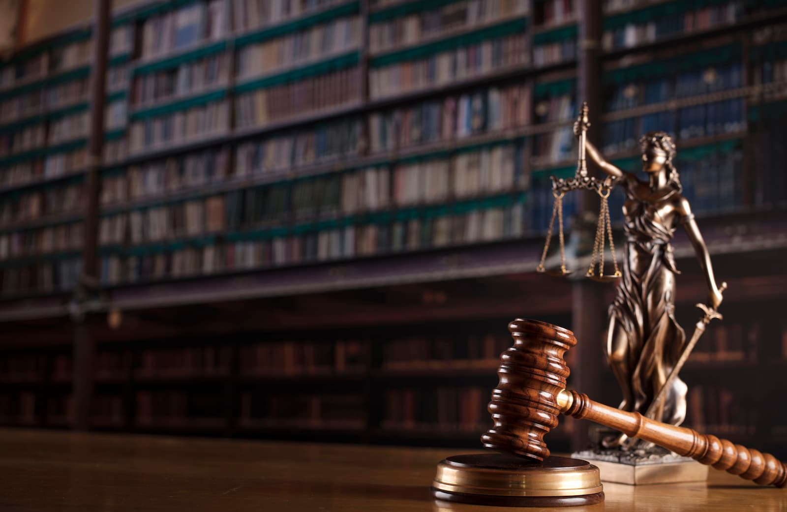 A wide, professional photo of a wooden desk in a law office. A wooden gavel and a bronze statue of Lady Justice are on the right side of the desk. The statue is holding a scale in one hand and a sword in the other. The background is a large, blurred bookshelf filled with legal books. The lighting is low and dramatic.