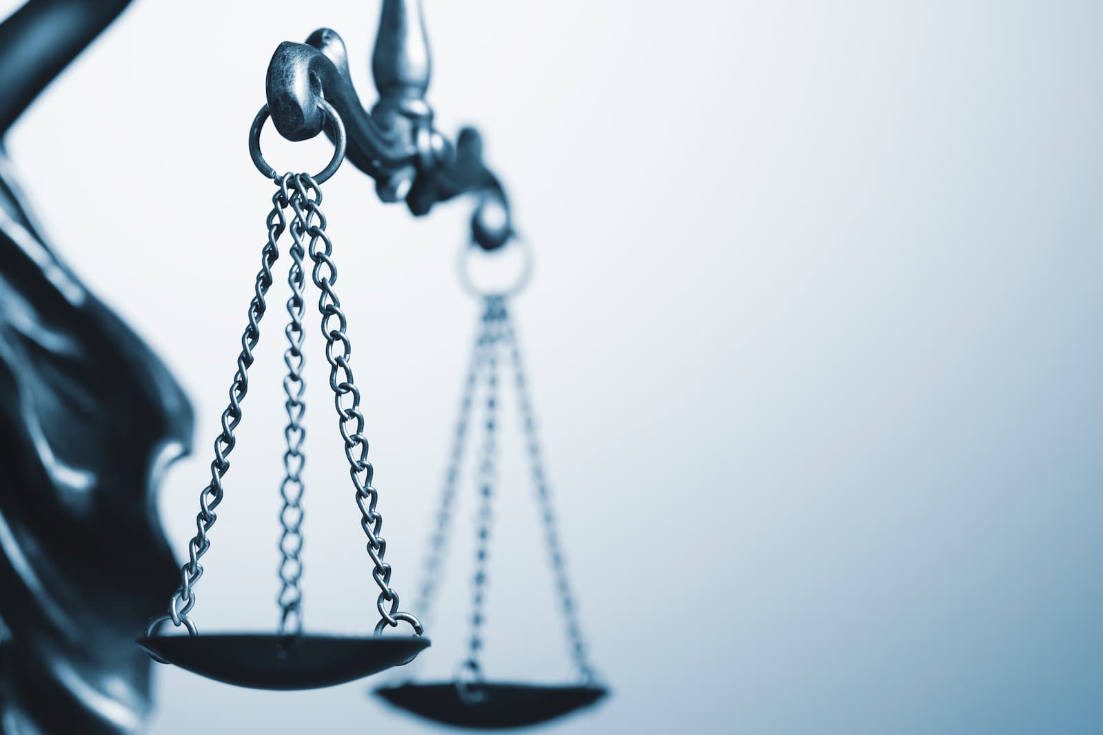 A close-up, low-angle photo of a scale of justice, with the focus on the left side of the scale. The scale's metal pans and chains are visible, with the chains hanging from a central beam that extends to the right. The background is a blurry, light blue and gray, with the other side of the scale visible but out of focus.