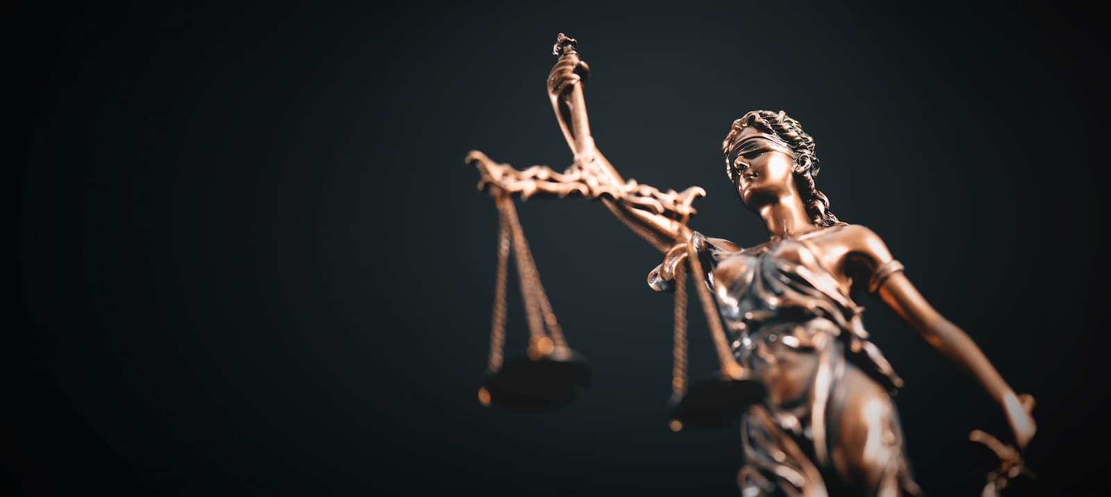 A dramatic, low-angle photo of a bronze statue of Lady Justice. She is blindfolded, holding a set of scales high in her raised left hand, with the scales hanging unevenly. Her right hand rests on the hilt of a sword. The statue is lit from the front, creating a bronze sheen, and is set against a plain black background.